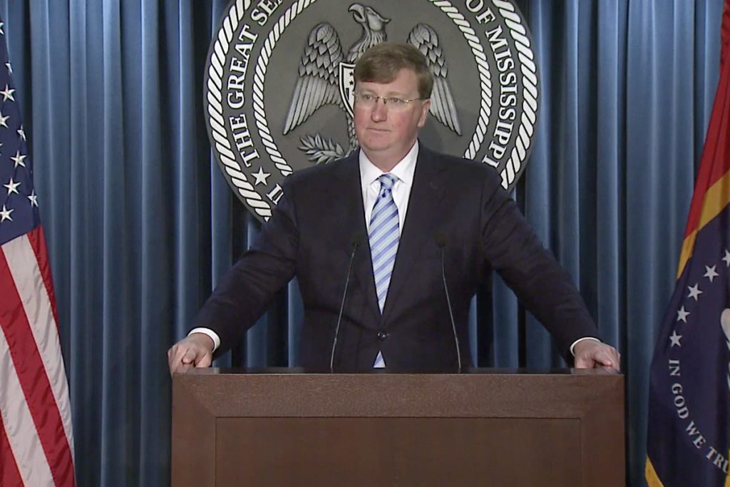 Tate Reeves standing at a podium in a black suit. The US flag is to his left and the MS flag to his right.