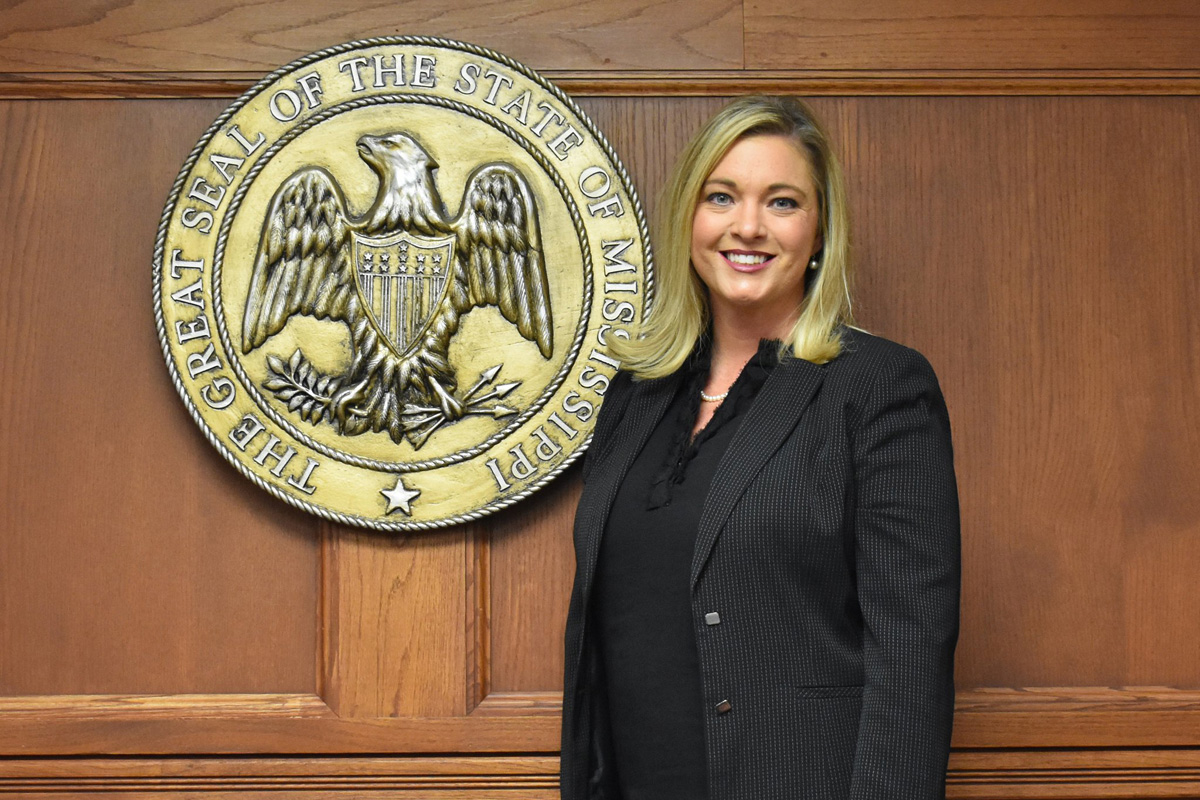 Tiffany Grove wearing a black suite with shoulder length blonde hair standing beside a golden decoration that says "The Great Seal of the State of Mississippi"