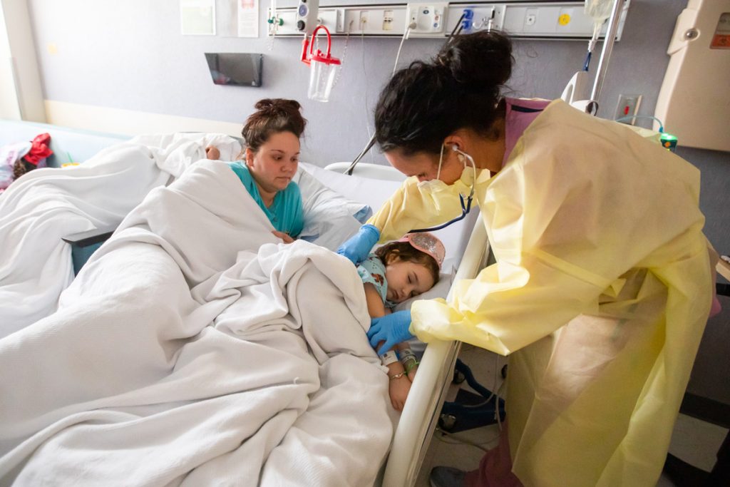 A mother and child in a hospital bed, as a doctor in yellow protective gear looks at the child