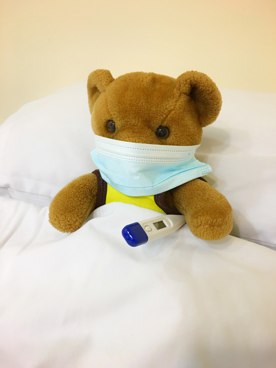 Brown teddy bear, tucked into bed. It's wearing a medical mask and has a thermometer tucked under one arm.