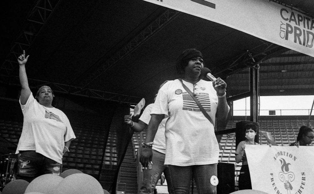 a photo of Valencia Robinson holding a microphone on a stage, Michelle Colon with her finger pointing skyward behind her