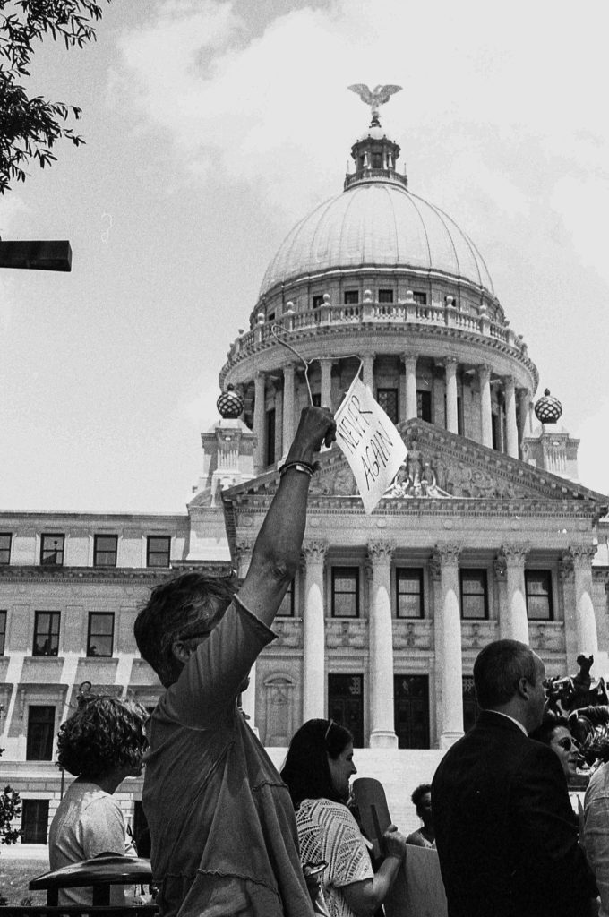a woman holds a coat hanger with the words "Never again" on it in front of the Mississippi Capitol