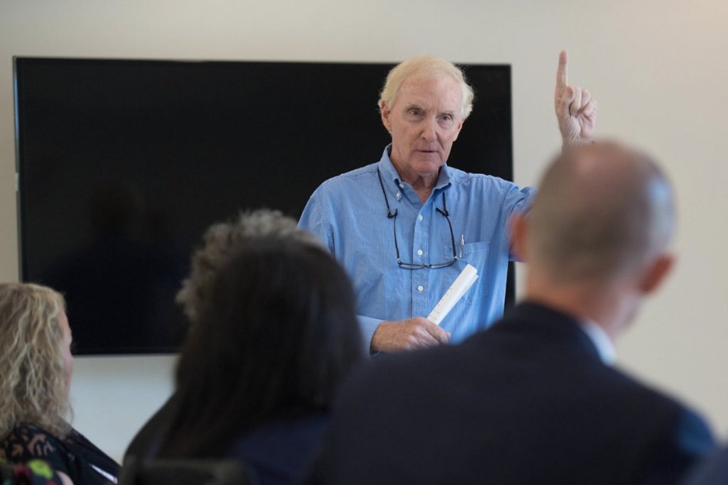 Andy Mullins in a blue button down shirt talking to a group of people with one hand pointing up