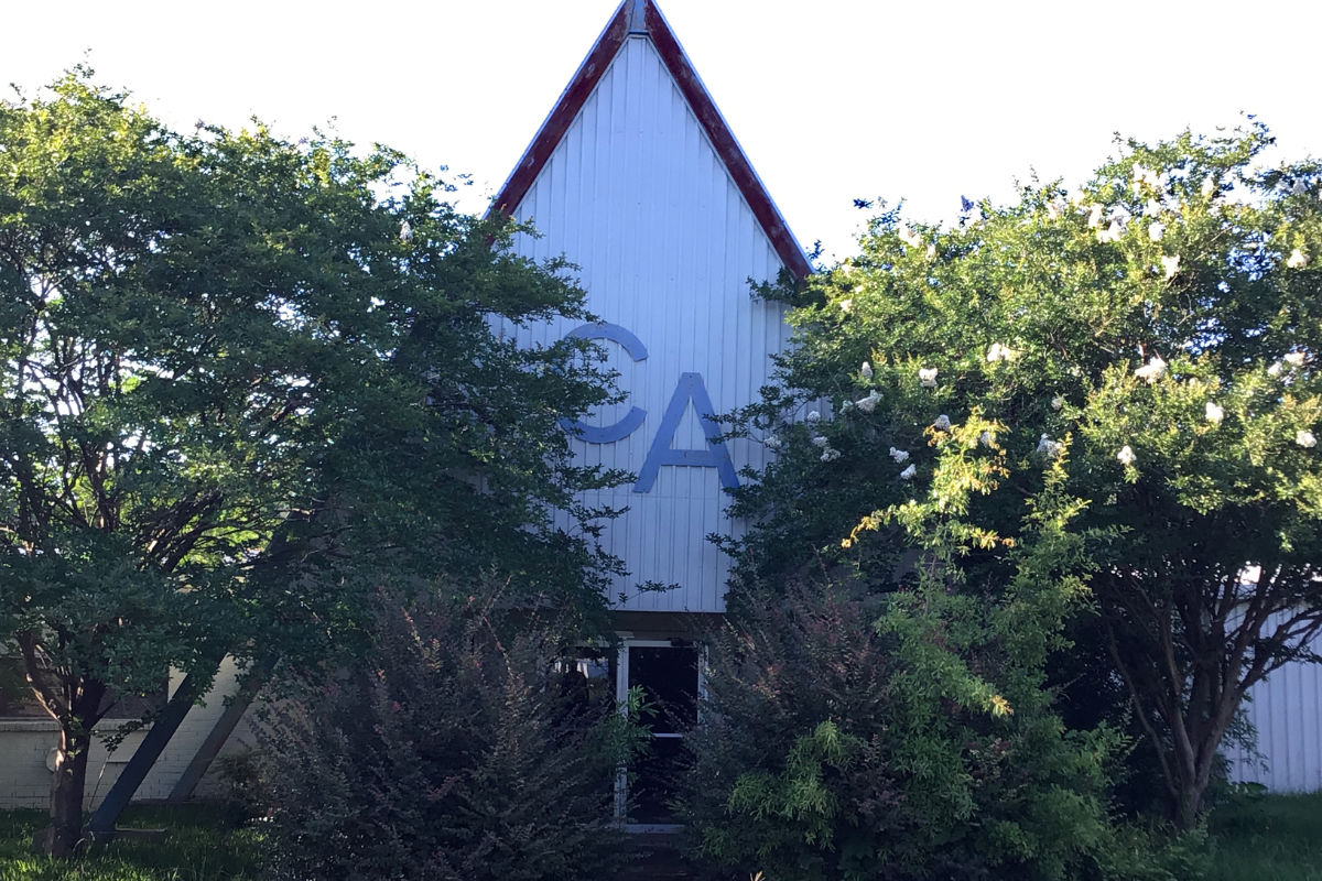 A white triangular building with large letters C A on the front over a doorway. Trees are overgrown in front.
