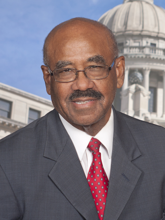 An older man with a mustache and glasses, wearing a dark grey suit and patterned red tie. The Mississippi Capitol is behind him