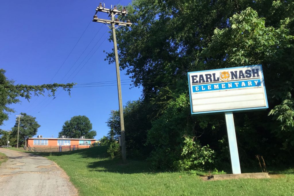 Earl Nash Elementary school sign in the foreground of a road on the left. A brick school building sits in the background behind green overgrowth