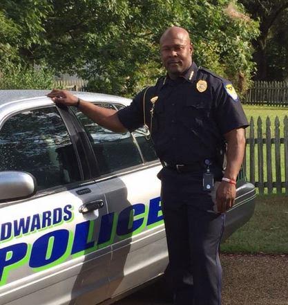 A bald man in a black police uniform rests a hand against a Edwards Police car