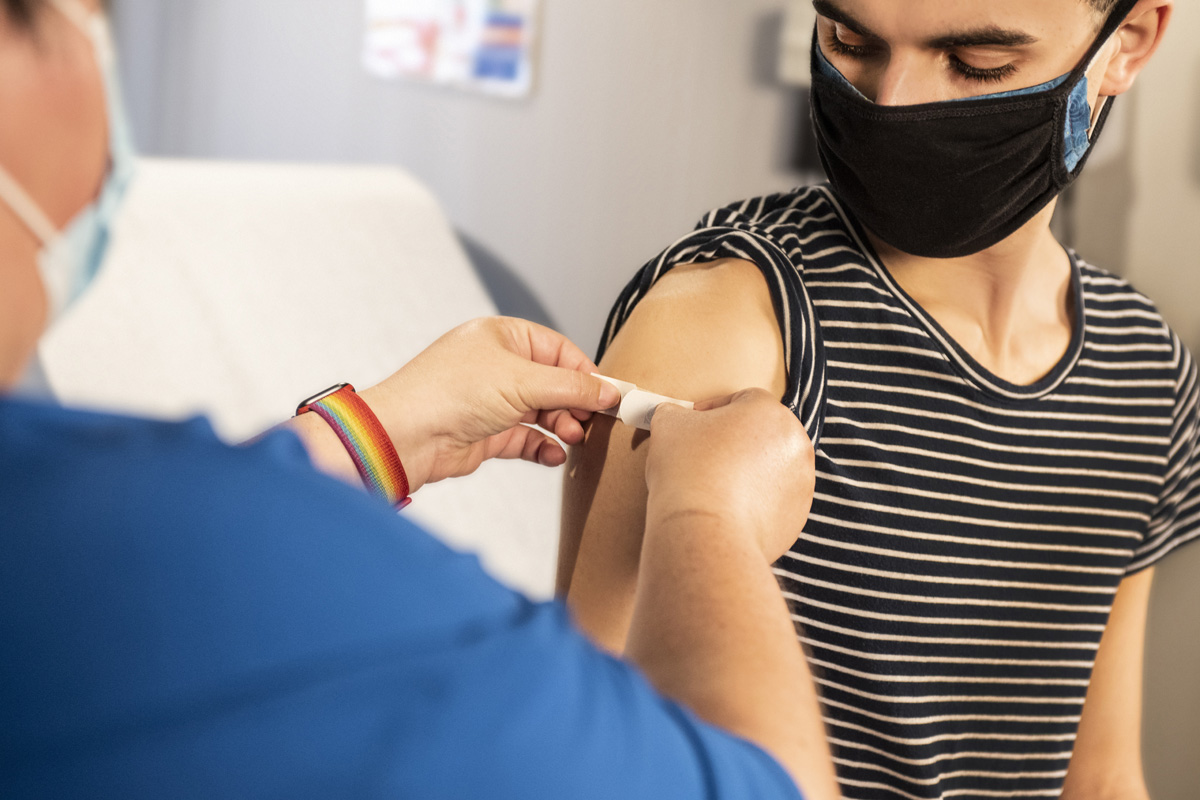 A masked person in a black and white stripped top to the right, a masked person in blue to the left putting a bandaid on the first persons arm