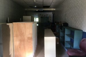 A jacket lies on empty bookcases stacked on their sides behind the glass doors of the John L. Barrett Elementary School. In the back, construction-paper cutouts circle a blackboard. A maroon office chair sits dusty near the glass doors.