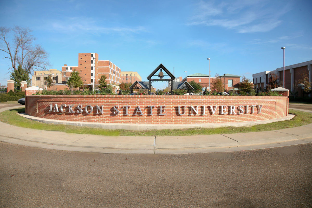 Jackson State University sign on a short red brick wall with the college campus behind it