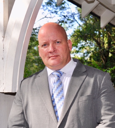 A bald man in a light grey suit and blue striped tie standing on a porch with trees behind him