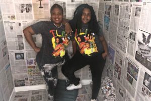 Two Black women wearing TLC tshirts pose in the center of a small room wallpapered in newspaper pages