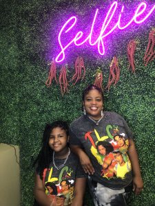 Two women wearing TLC tshirts pose in front of a green ivy wall with a pink neon sign that says Selfie