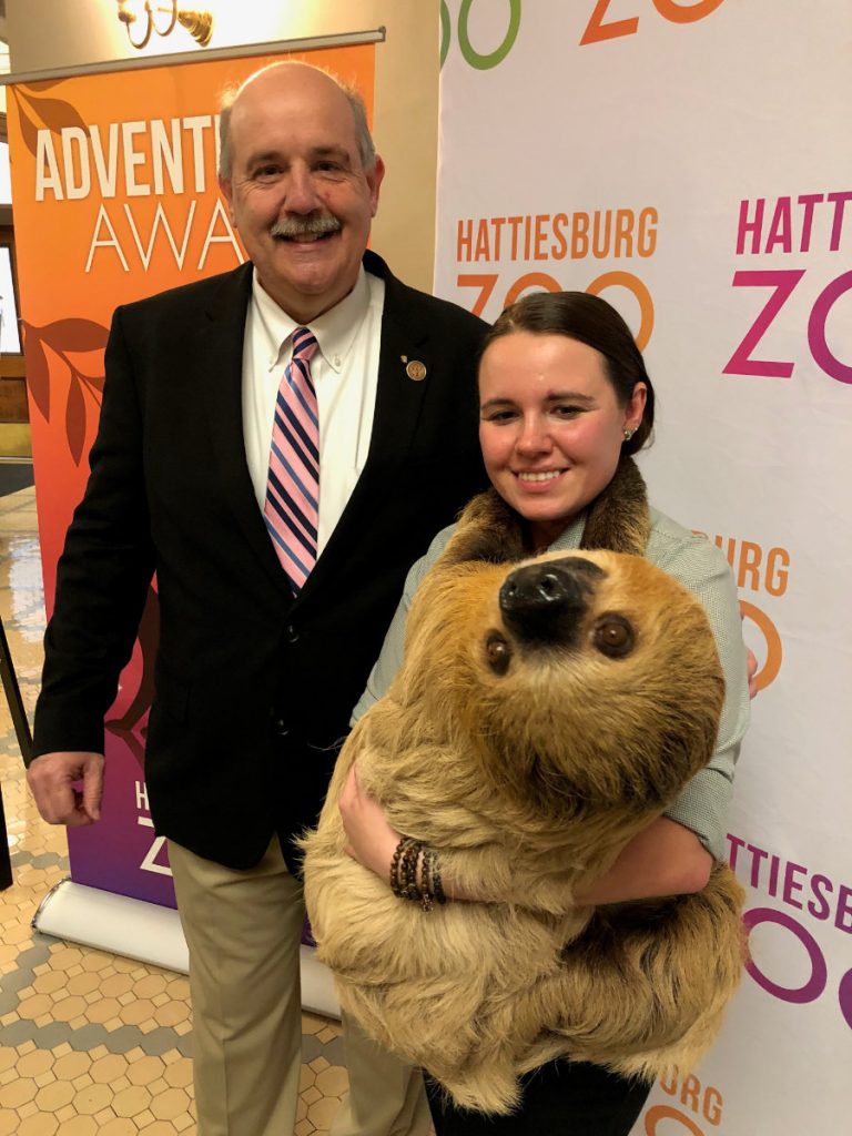 An older man in a suit stands beside a younger shorter woman. The woman is holding a large sloth that's looking at the camera upside down