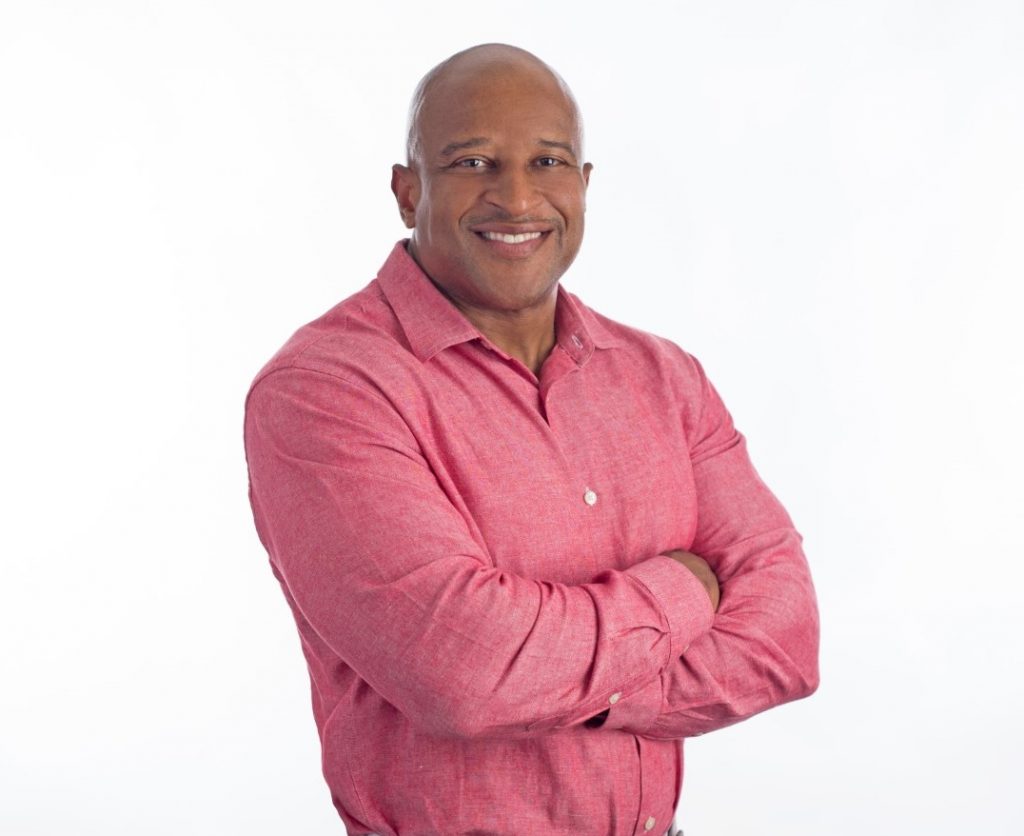 A smiling bald man wearing a pink button down shirt with his arms crossed