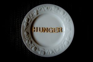 A white china plate with fruit motif embossed on the rim, with brown wooden letters that spell out HUNGER in the center of the plate