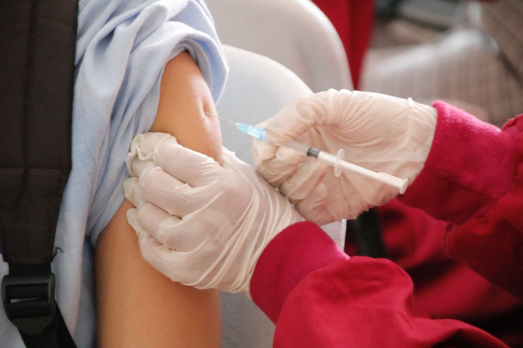 To the left is a person with a light blue rolled up sleeve, to the right is a person with long red sleeves and gloved hands administering a vaccine