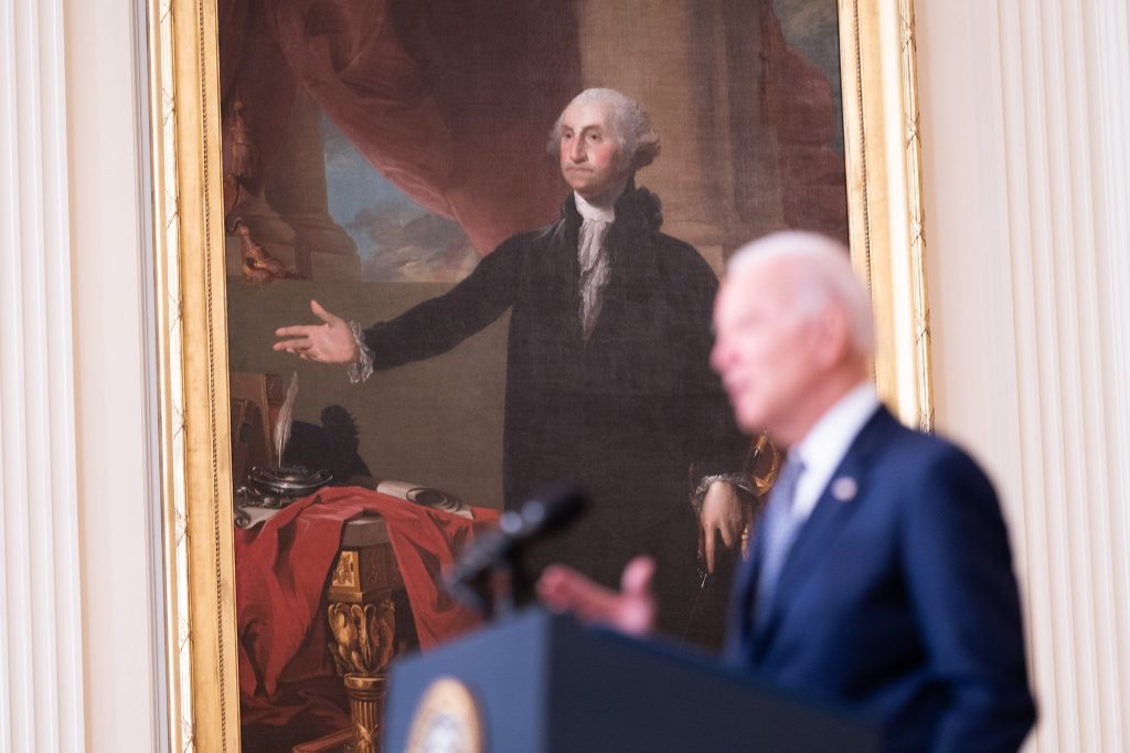 A photo of President Joe Biden speaking in front of a portrait of George Washington