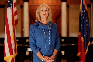a photo of Lynn Fitch standing between a Mississippi and US flag. She is suing stop Biden's vaccine mandate