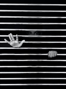 Black and White photo of a man looking through horizontal bars, with his hands sticking through