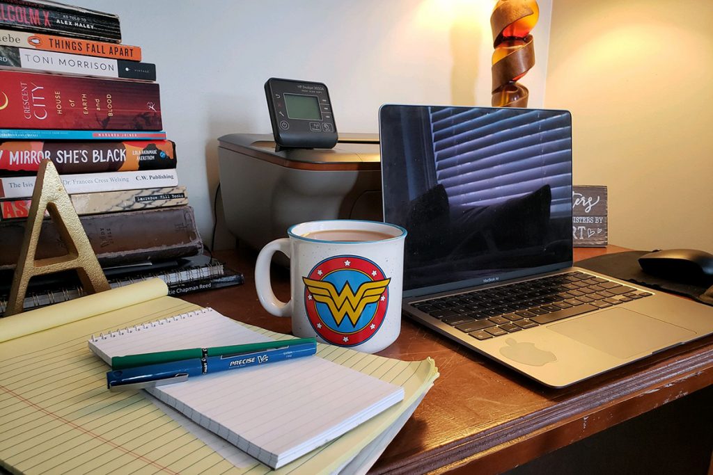 Desk with laptop, notepad and pen, stack of books and cup of Wonder Woman coffee