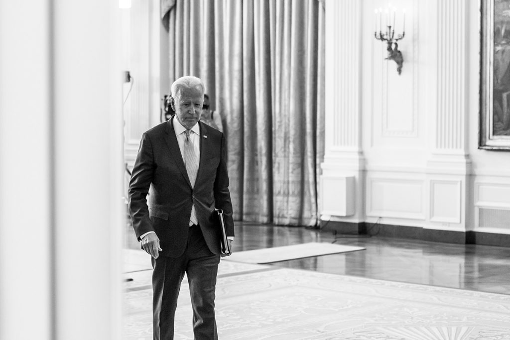 Joe Biden walks with folder under arm though a White House hallway