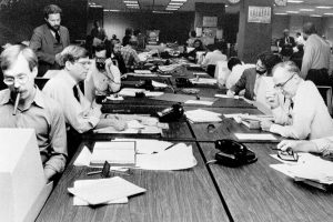 Black and white photo of white men in button up shirts sitting around a desk filled with papers and phones
