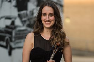 Shelly Kemp, long brown hair worn down and a sleeveless black dress, holding a violin