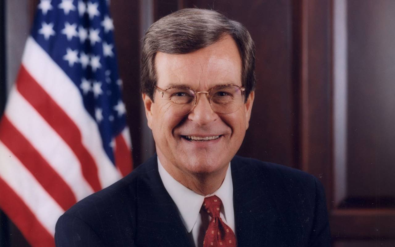 Headshot of Trent Lott in a suit and tie wearing glasses. A wooden wall with a US flag is behind him.