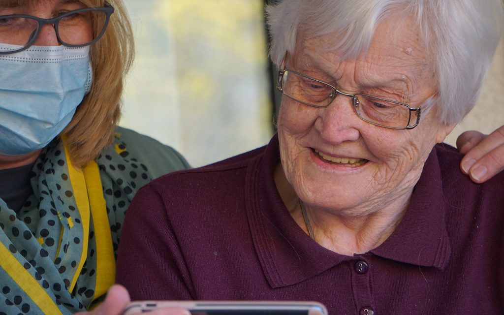 A grandmother in her nineties getting to see her grandchildren via mobile device as a masked family member holds the phone