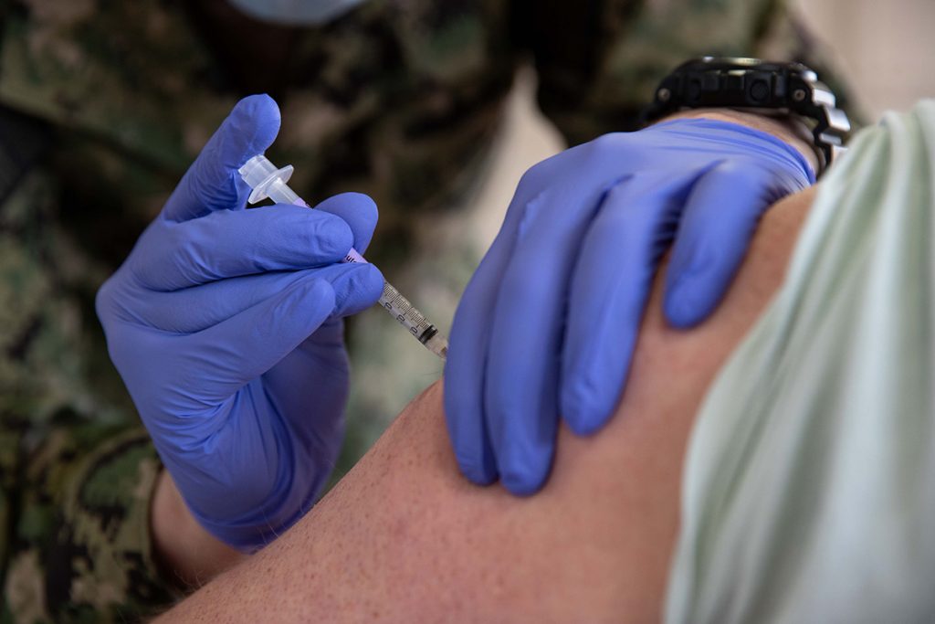 Blue gloved hands administer a vaccine in the arm of a person with rolled up sleeve