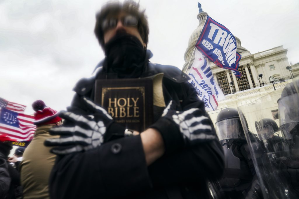 a photo of a man with skeletal gloves holding a Bible outside the U.S. Capitol as Trump flags and American flags fly behind him
