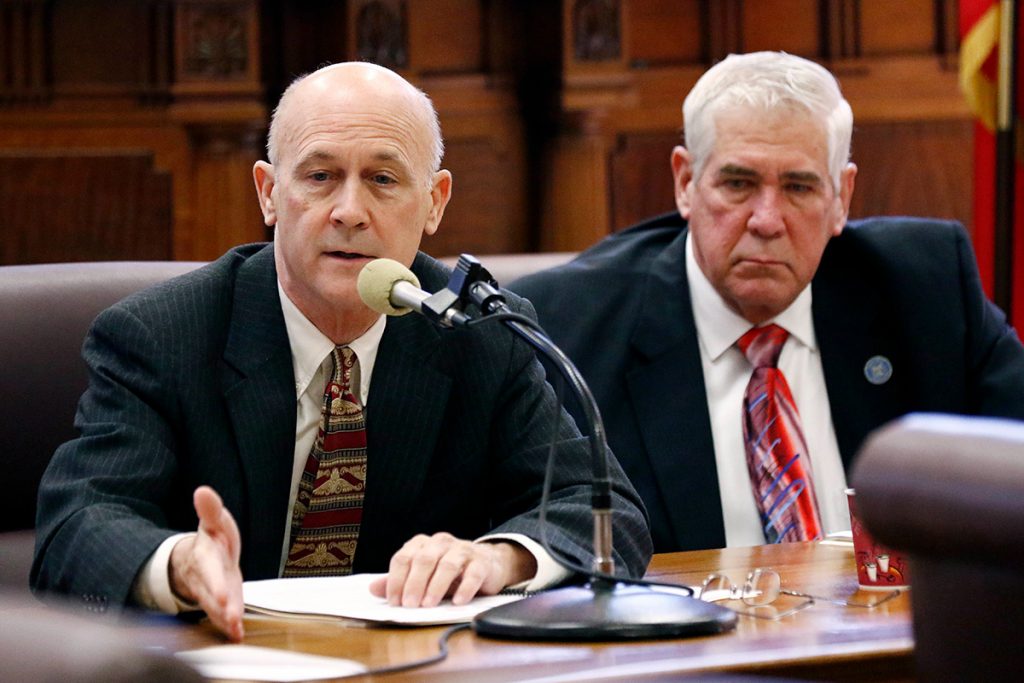 Two older men in dark suits sit in legislature at a mic