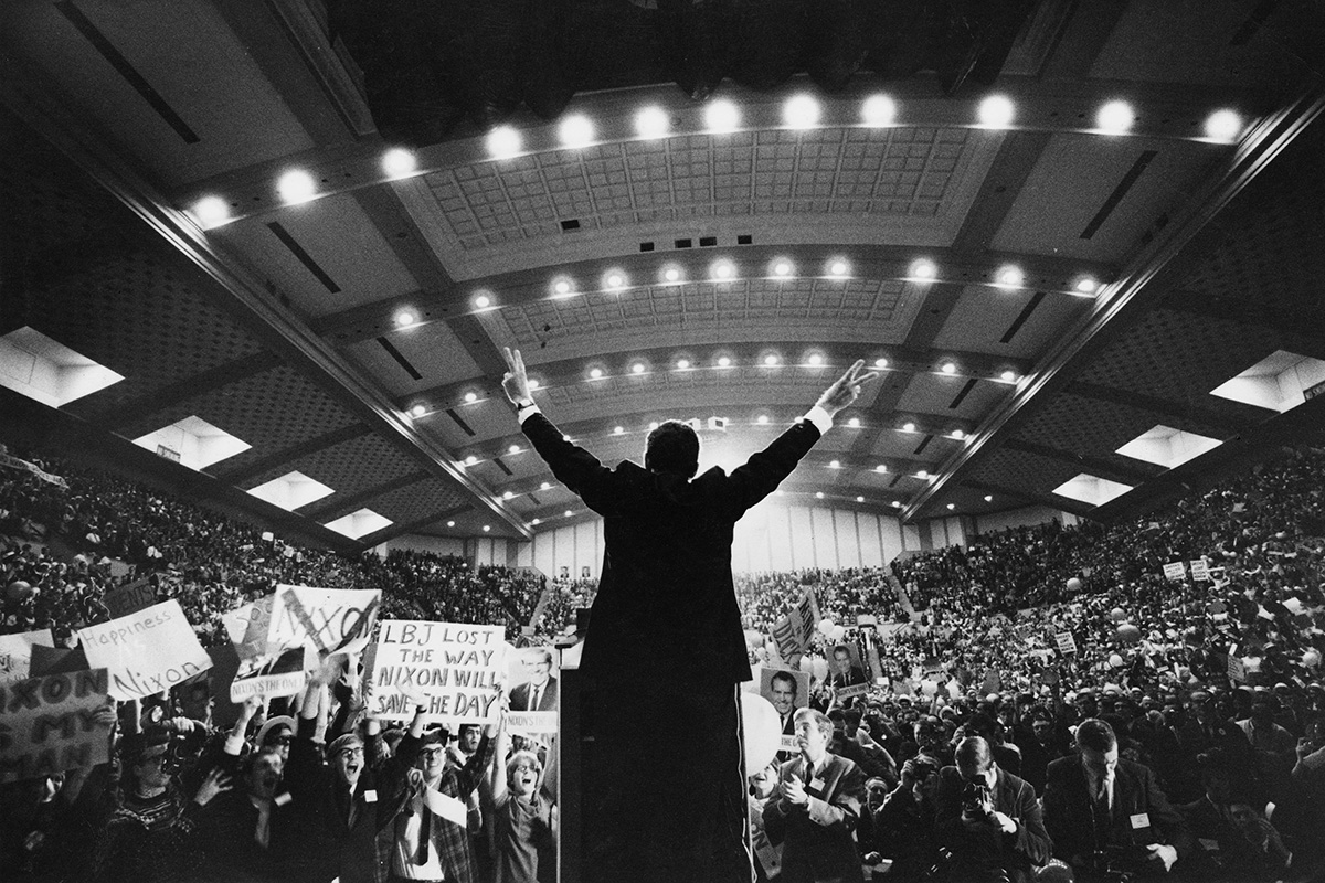 Richard Nixon, in a dark suit and flashing a 'V for victory' sign with both hands in front of an arena of supporters.