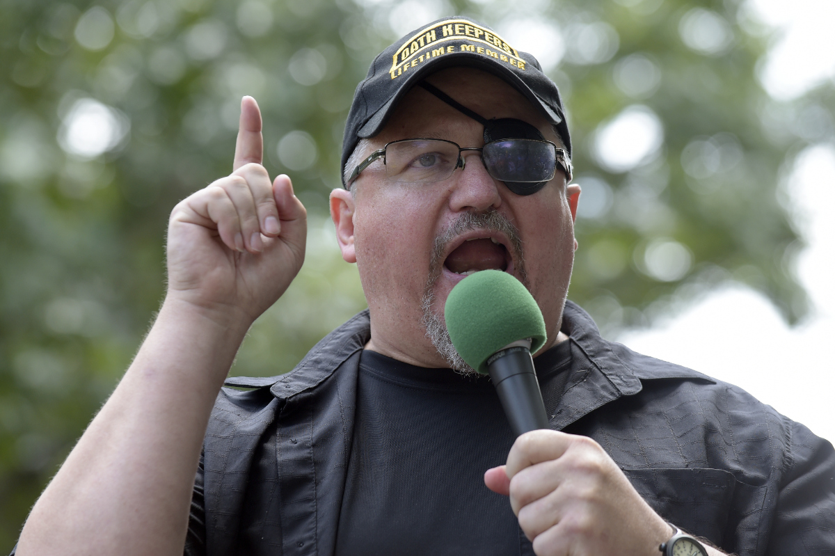 White man wearing an Oath Keepers cap, glasses, eye patch speaking into a microphone with one hand pointed upward