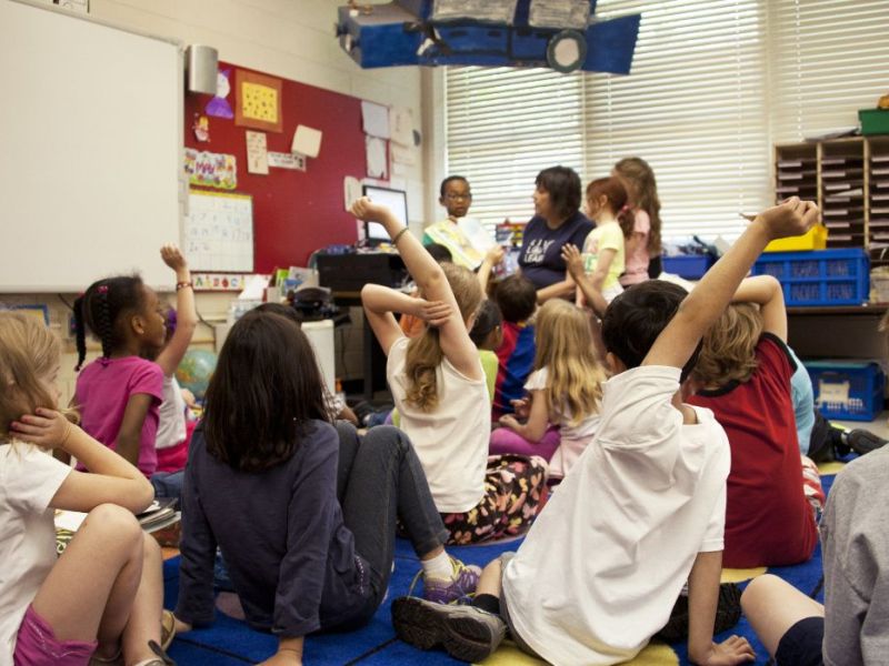 Several children seated on a carpet in a classroom with a teacher at the front of the group.
