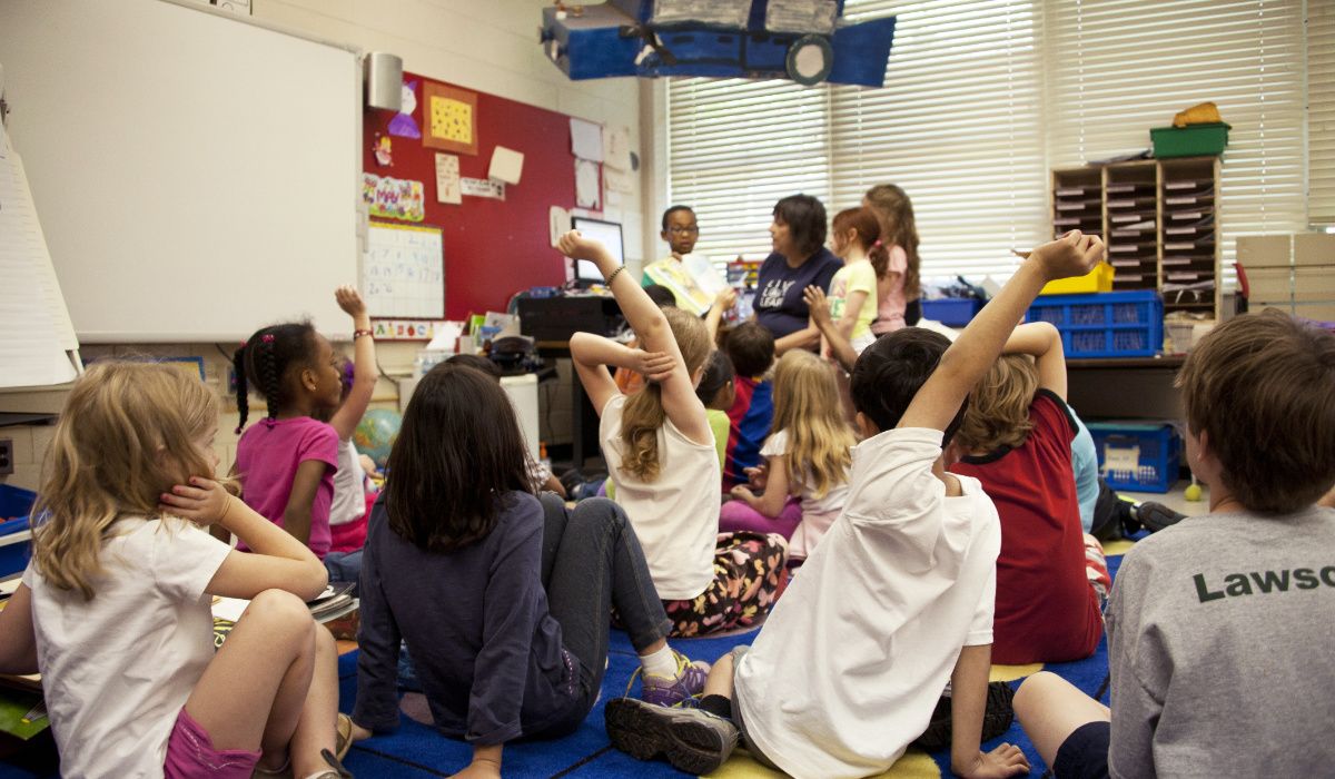 Several children seated on a carpet in a classroom with a teacher at the front of the group.