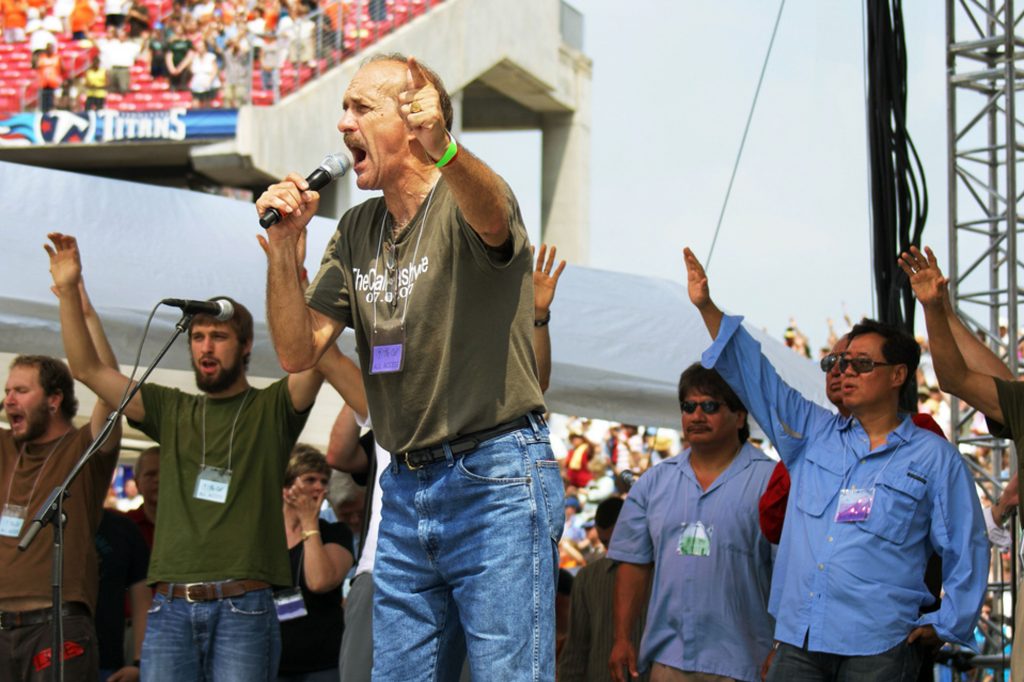 A photo of Lou Engle speaking at a stadium outdoors, men behind him with their arms lifted in prayer