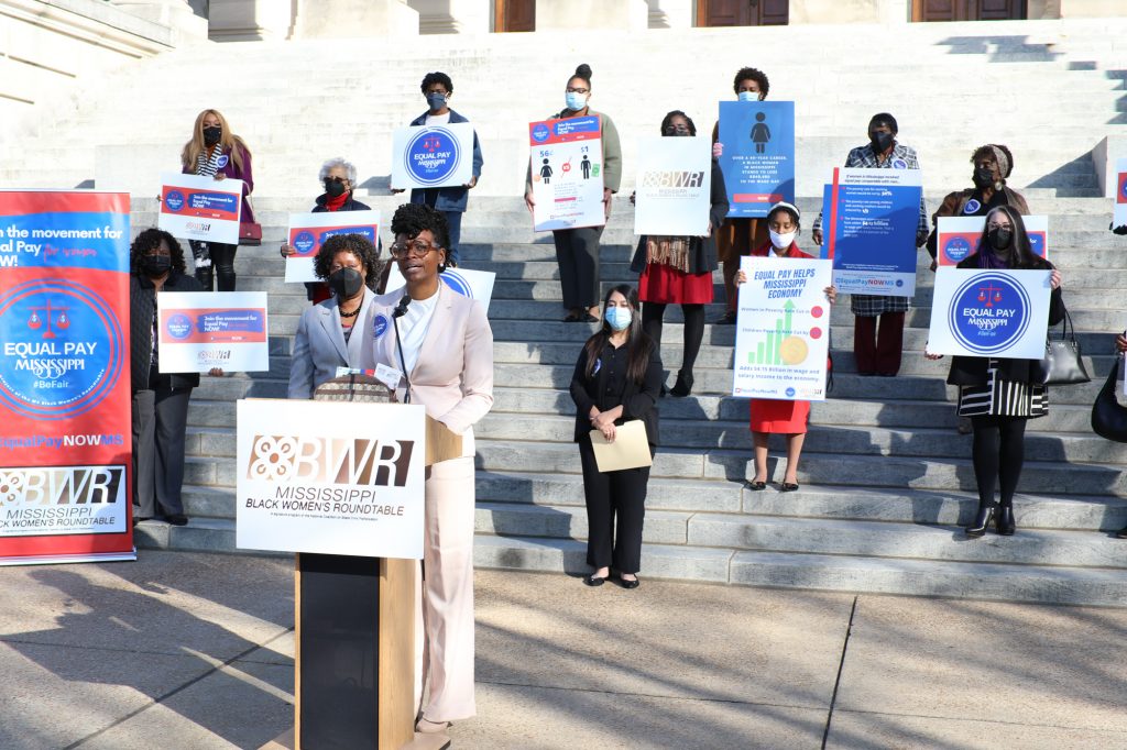a photo of Cassandra Welchlin speaking outside the steps of the Mississippi Capitol with people holding equal pay signs behind her