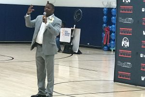 Black man in a suit talking to crowd with a microphone in a new gym in Brooklyn, N.Y.