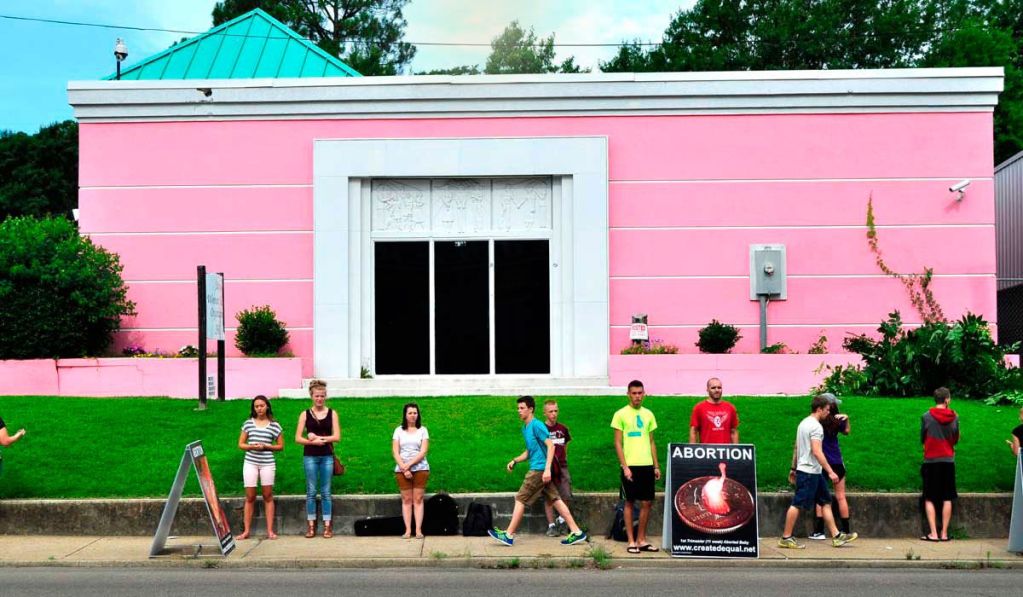 People stand in a line outside a pink building.
