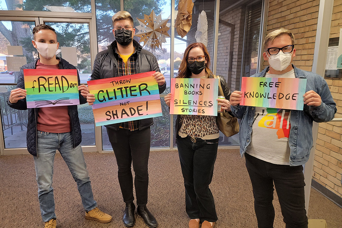 Four masked activists holding rainbow signs "fREADom" "Throw GLITTER not SHADE!" "Banning Books SILENCES Stories" "Free Knowledge"