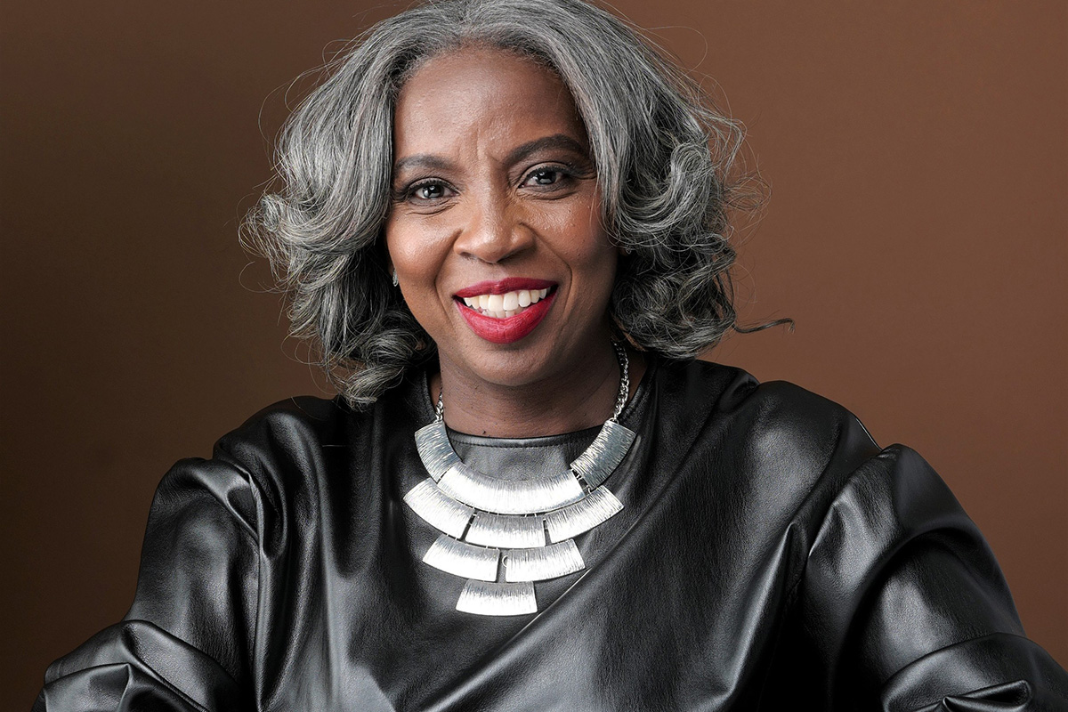 Woman in black long sleeve top that looks like smooth leather, a bold silver necklace, and silver hair that frames her face. She's sitting against a brown background