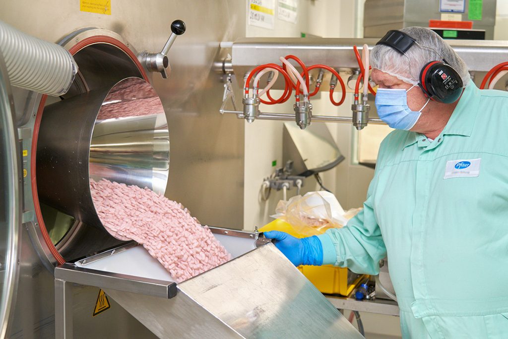 A man in a mint green Pfizer jumpsuit overlooking pink pills pouring out of a machine