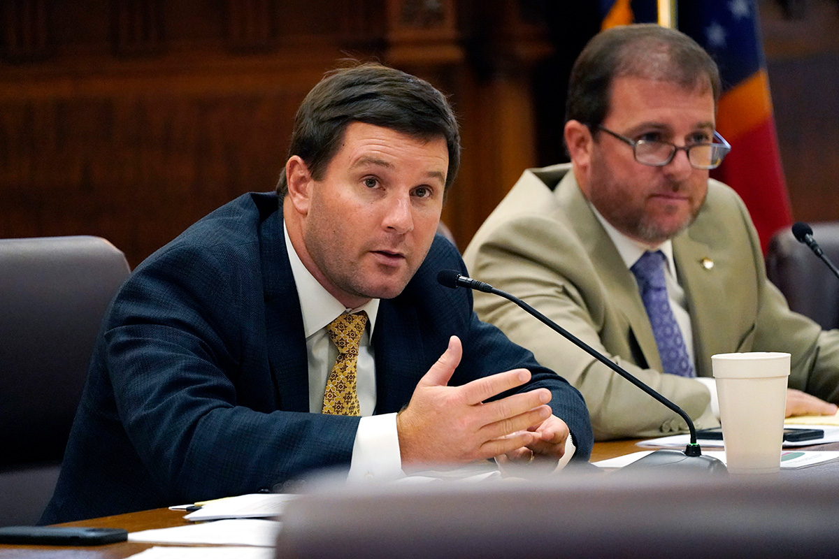 Two men in suits sitting at a desk talking into thin mics