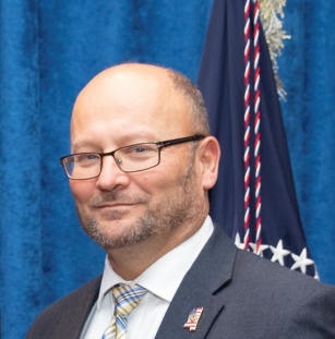 Man with glasses in a suit and plaid tie with a Mississippi lapen pin