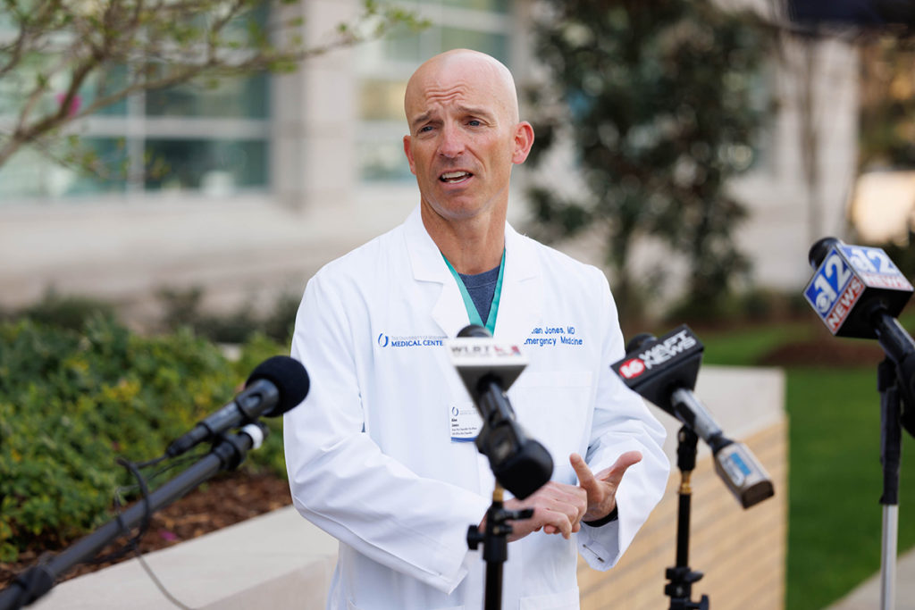 A man in a white medical coat speaks outside in front of four microphones
