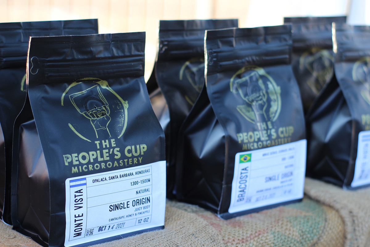A row of black coffee bags on a table covered in burlap cloth, the bags say "The People's Cup Microroastery"