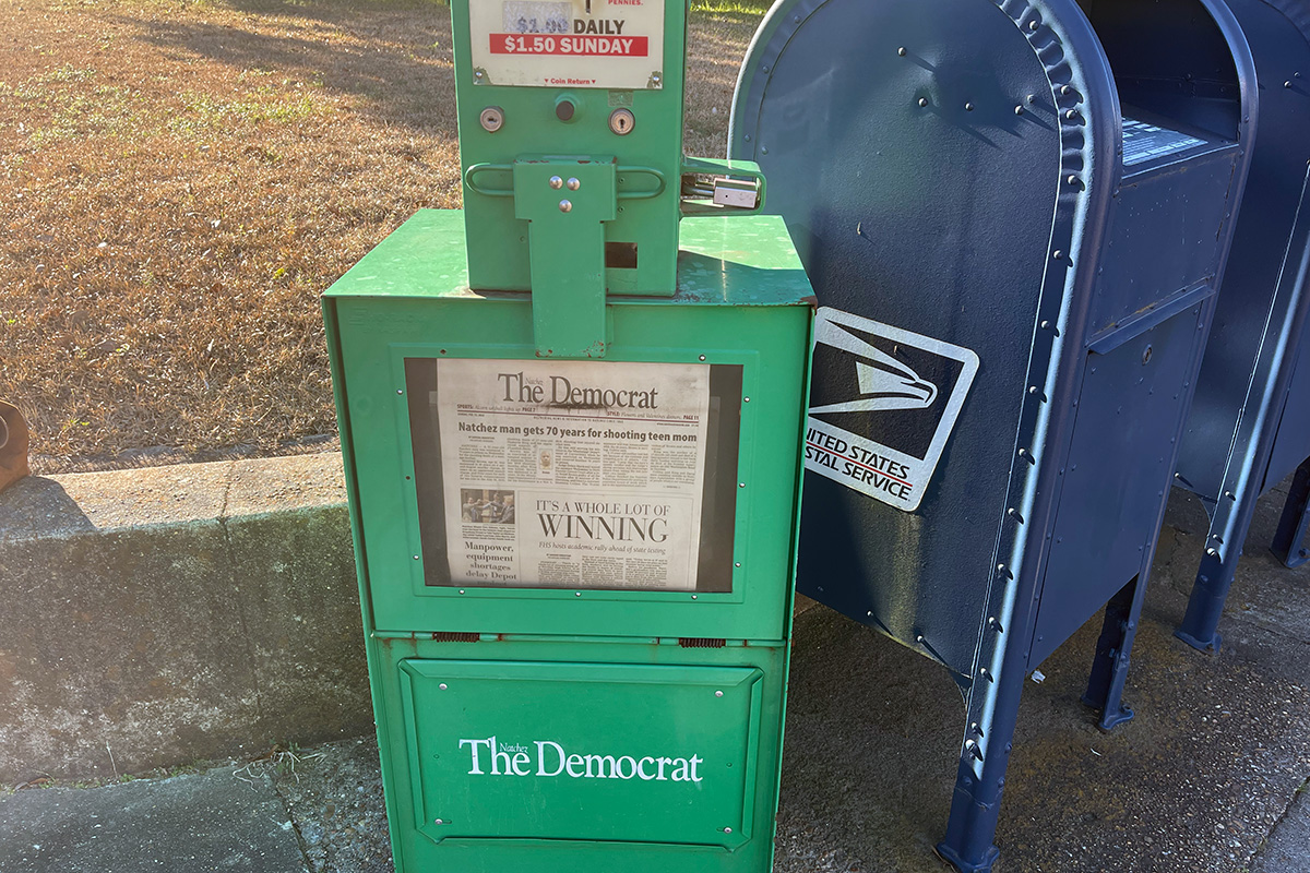 A green newspaper stand for The Nachez Democrat, beside blue mailboxes