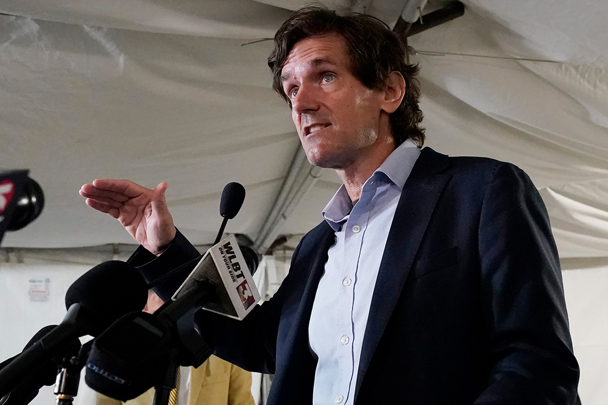 A man in a dark suit and light blue button up shirt open at the collar, speaking in from of microphones.
